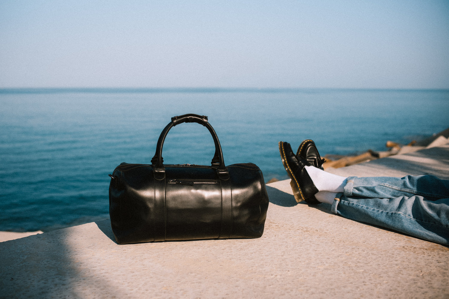Schwarze Weekender-Tasche aus Leder von Buckle & Seam – stilvoll platziert am Meer, ideal für stilvolle Kurztrips und Reisen